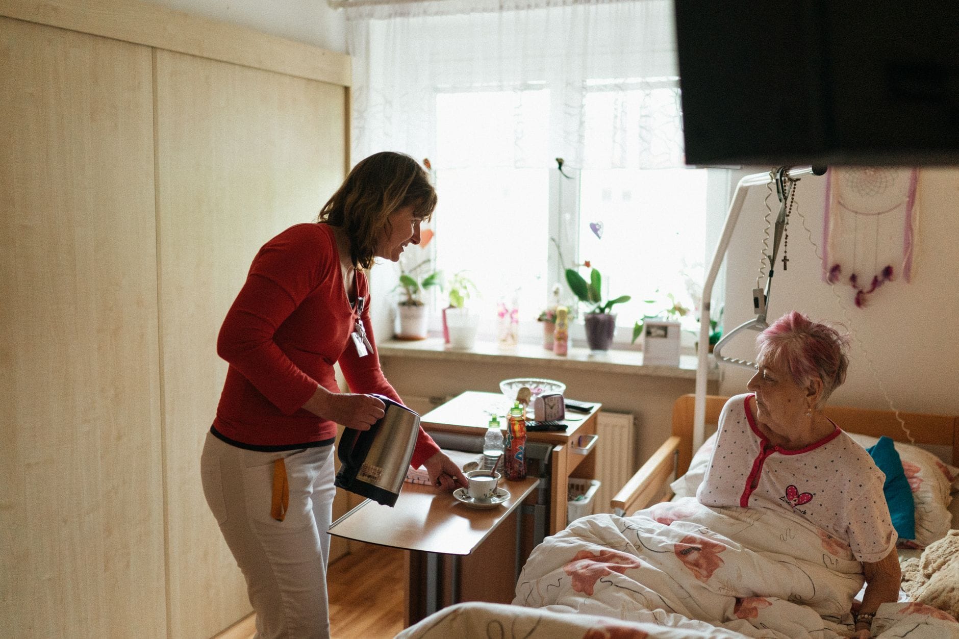Opiekun podaje herbatę seniorce w przytulnej sypialni, troskliwa opieka